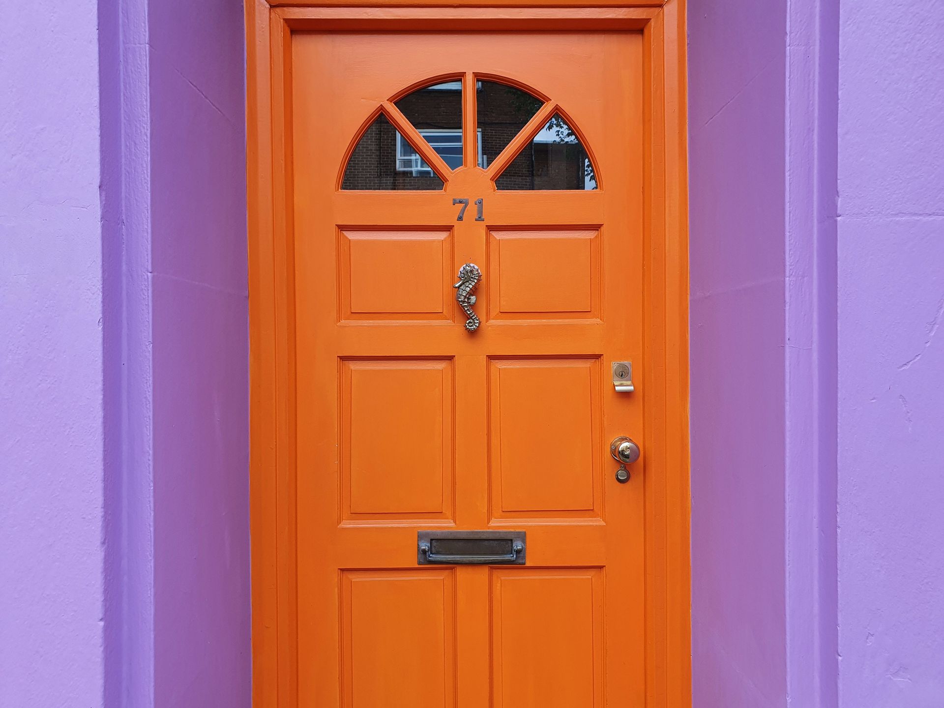 A striking orange door with a seahorse knocker against a purple wall, numbered 71.